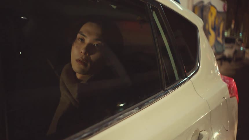 A contemplative Asian man rides in the backseat of a car at night, watching the city lights reflect in the window