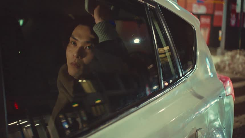 A pensive young Asian man looks out the window of a moving car at night as the city lights reflect in the glass