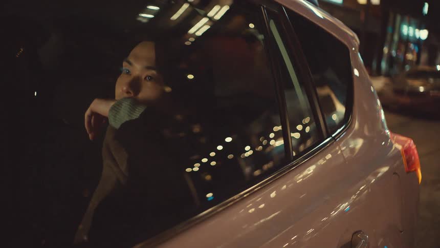 A thoughtful young man sits in the back of a car at night, gazing out the window as the city lights reflect on the glass