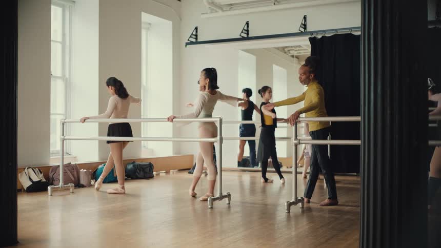 A male ballet instructor teaches a group of young dancers proper form at the barre in a sunlit studio