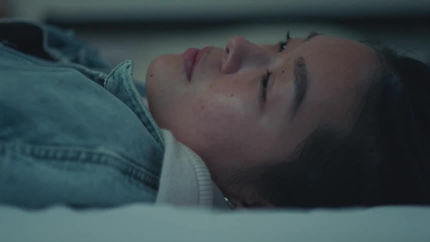 A tired and pensive young Asian woman lies down on her bed with her eyes closed, finding a moment of rest