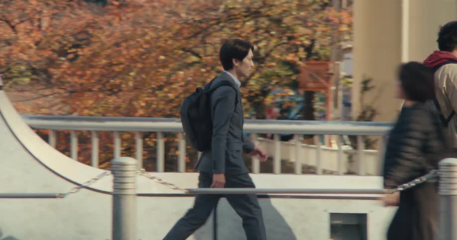 A young Japanese businessman wearing a suit and backpack walks across a city bridge on his commute during a sunny autumn day
