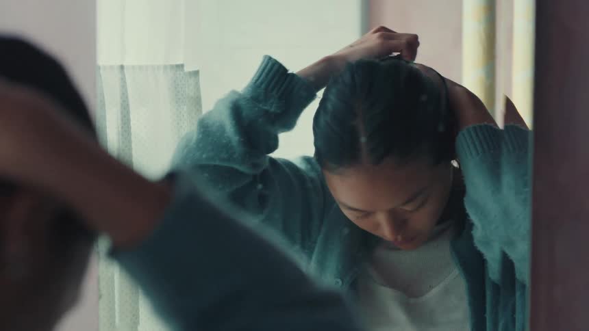 An introspective young woman with a somber expression ties her dark hair into a ponytail while looking at her reflection in the bathroom mirror