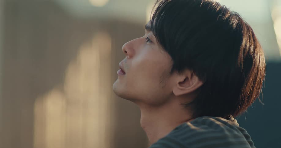 A thoughtful young Asian man sits in quiet contemplation, bathed in the soft light of the afternoon sun