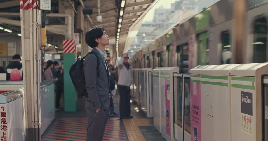 A young Japanese businessman in a suit patiently waits on the platform as his train arrives at the station