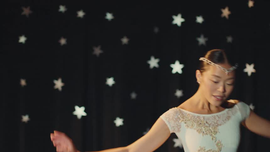 A graceful Asian ballerina in a white tutu performs an elegant slow-motion dance on a stage with a starry background