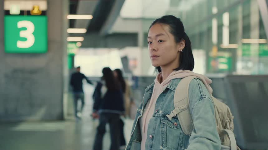A young Asian woman wearing a backpack and denim jacket walks through a modern airport terminal in slow motion