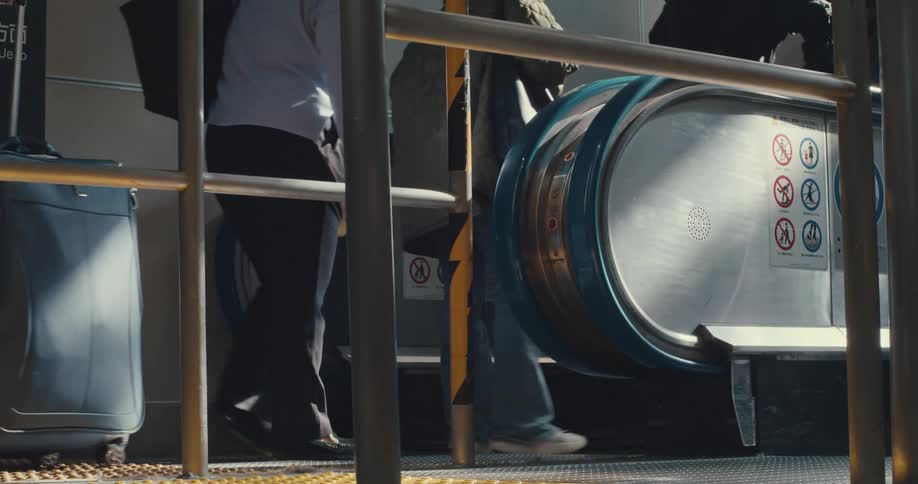 A slow-motion, low-angle view of commuters' feet and luggage as they step onto an escalator in a public transit station