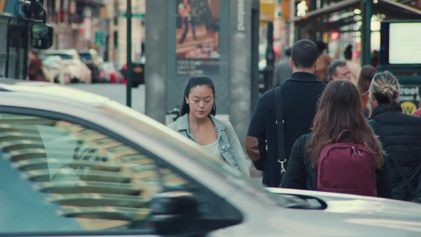 A young Asian woman walks toward the camera in slow motion on a busy city street sidewalk