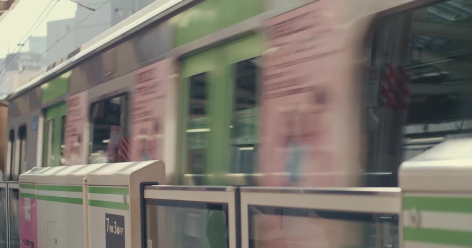 A close-up side view of a speeding green and silver commuter train as it passes a station platform in Tokyo