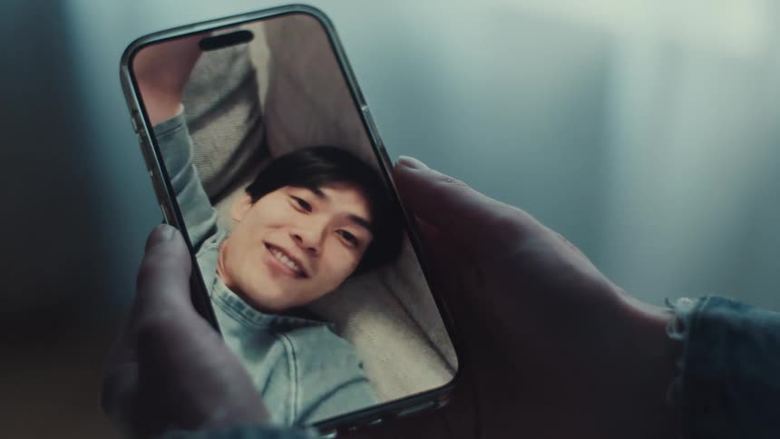 Close-up of a person's hands holding a smartphone during a video call with a smiling young man relaxing on a couch