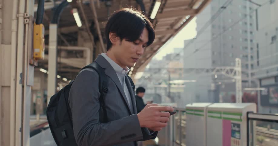 Young Asian businessman checks his phone on the train platform as he waits for his daily commute