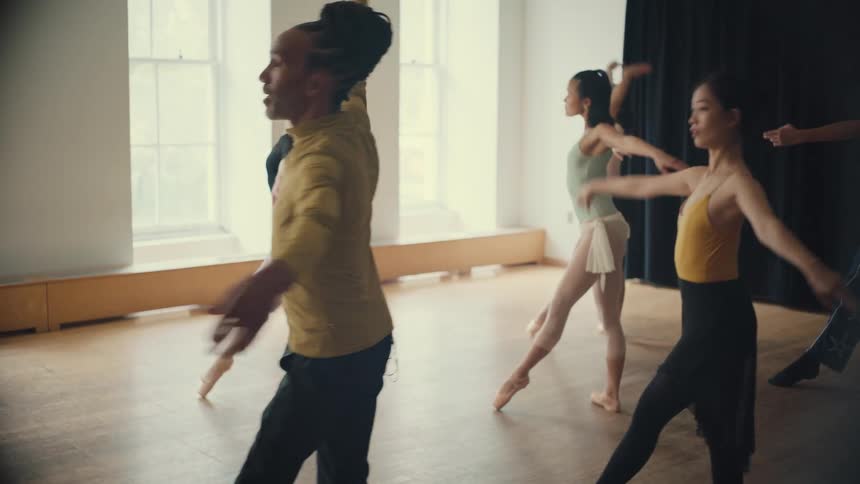 An instructor leads a diverse group of ballet dancers through a graceful routine in slow motion in a bright studio