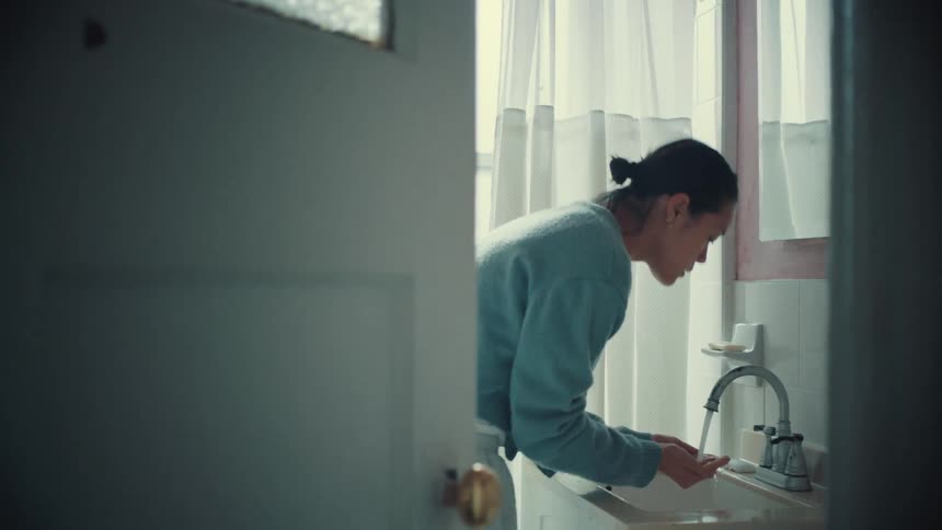 A woman seen through a doorway performs her morning routine, slowly tying her hair back and washing her face in the bathroom