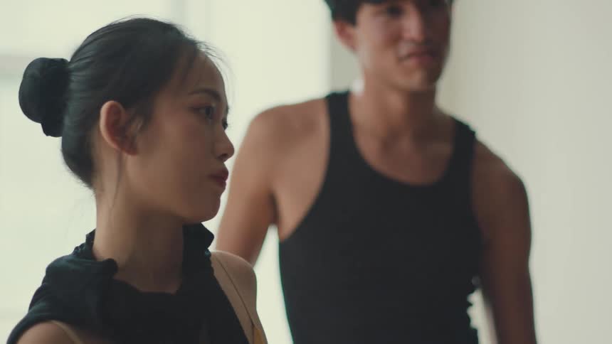 A focused young Asian ballerina intently listens to her colleague during a dance rehearsal in a bright studio