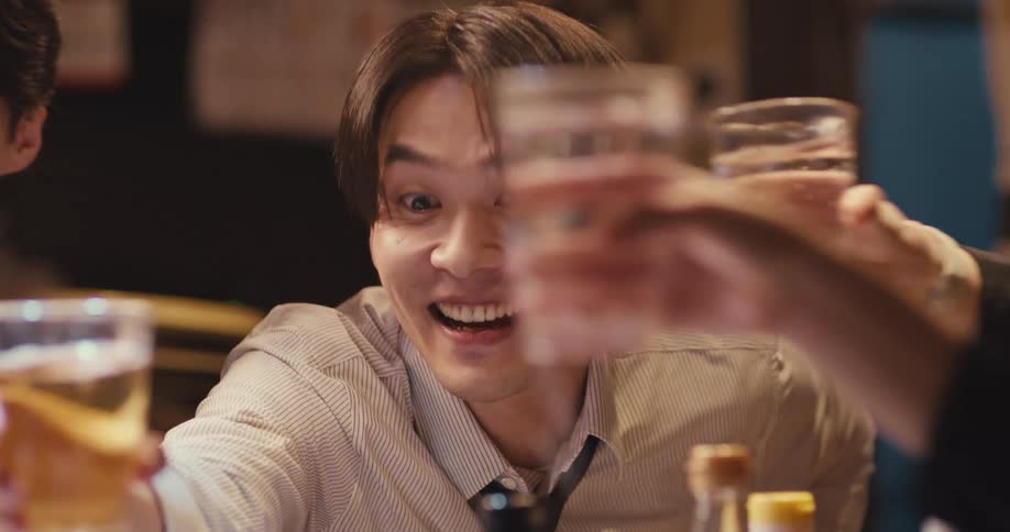 A cheerful young Asian businessman toasts with his colleagues at a bar after work, smiling as they clink glasses in slow motion