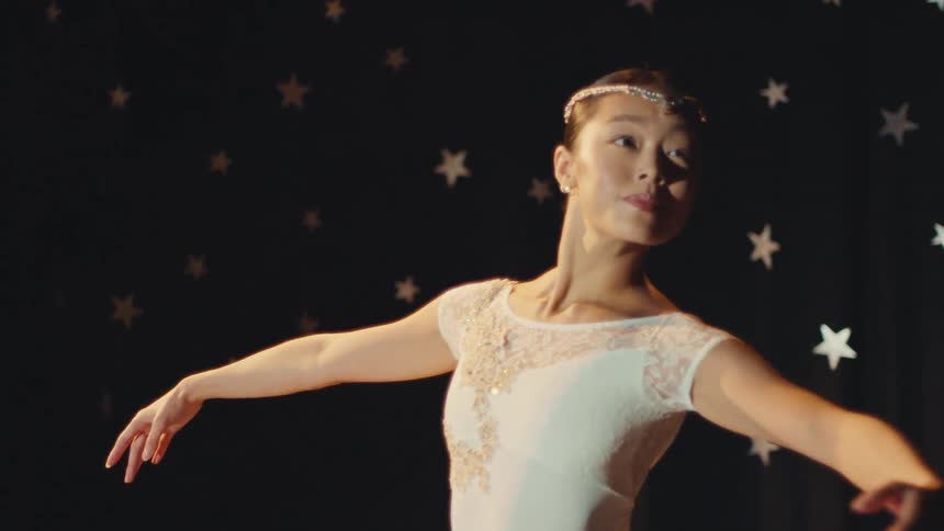 Slow motion close-up of a beautiful ballerina in a white costume gracefully performing on stage against a starry backdrop