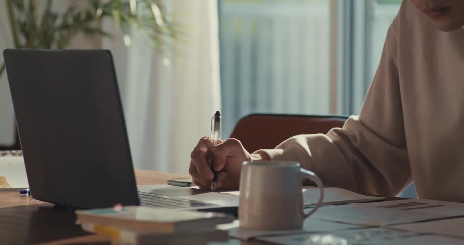 A person diligently takes notes while working on their laptop at a sunlit desk in a cozy home office setting