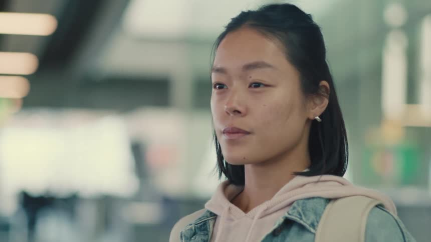 A thoughtful young Asian woman with a backpack waits patiently while standing in an out-of-focus terminal