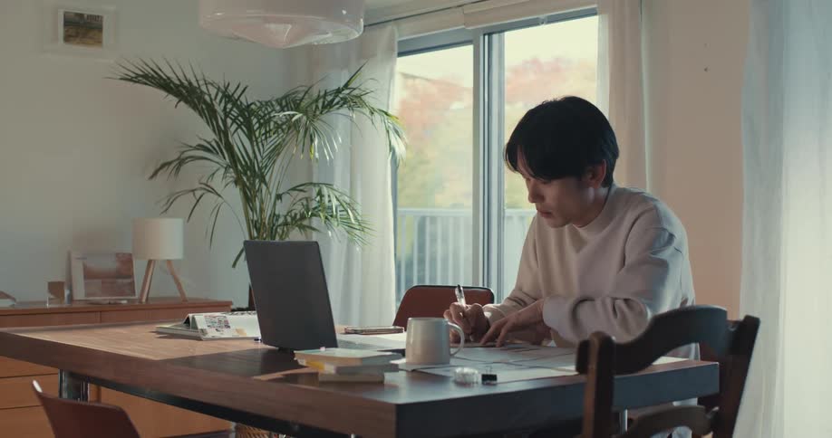 A focused young Asian man diligently works from home, writing at a sunlit dining table with his laptop nearby as the camera slowly pulls back