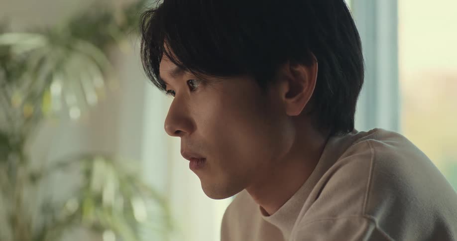 A close-up shot of a young Asian man looking down with intense focus and concentration in a sunlit room