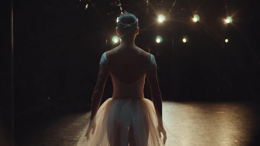 A graceful ballerina in a white tutu walks onto a dark, spotlit stage, seen from behind in slow motion