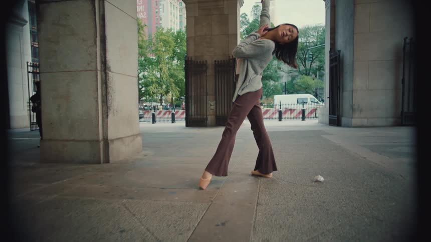 A graceful young Asian ballerina performs an expressive contemporary dance in slow motion under a grand city archway
