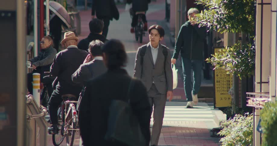 An Asian businessman walks purposefully through a busy, sunlit alleyway in a Japanese city