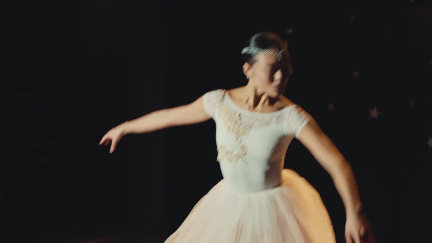 Slow-motion close-up of a beautiful ballerina in a white tutu gracefully performing a solo dance on a dark, spotlit stage