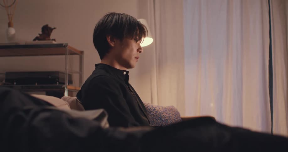 A pensive young Asian man sits alone on a sofa in his dimly lit apartment at night