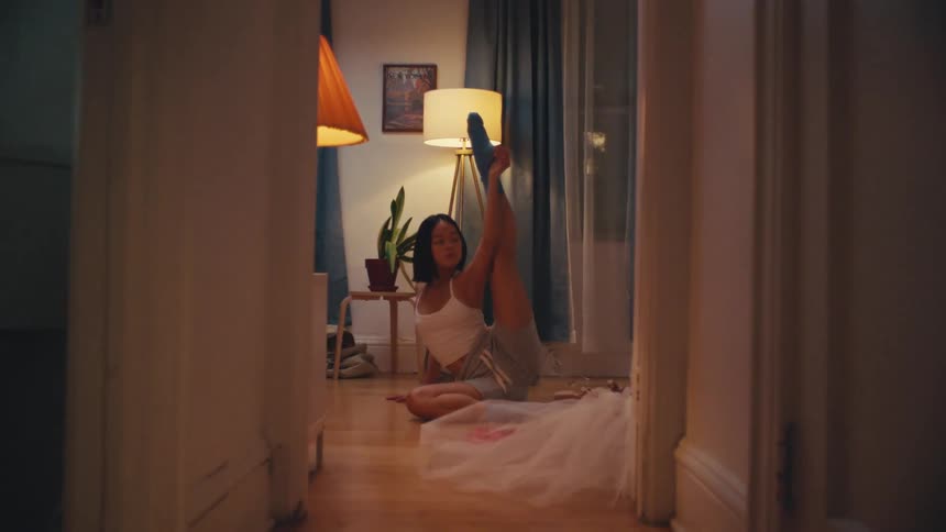 A dedicated young ballerina practices a difficult leg stretch on the floor of her cozy apartment at night
