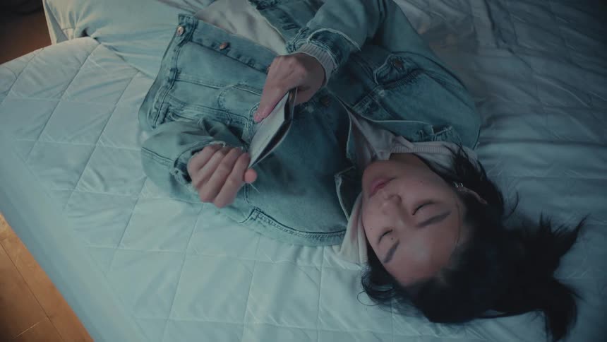 A tired young woman wearing a denim jacket and hoodie reads a book while lying on her bed before falling asleep
