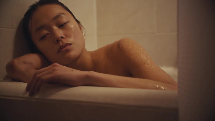 A young woman with a somber expression rests in a dimly lit bathtub, lost in thought