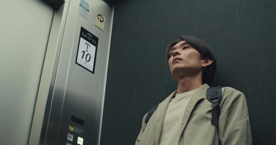 A pensive young man wearing a backpack stands in a moving elevator as the floor numbers ascend