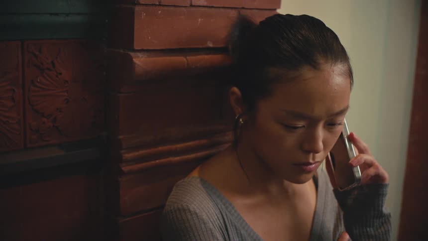 A sad young Asian woman listens to a difficult phone call in a dimly lit room
