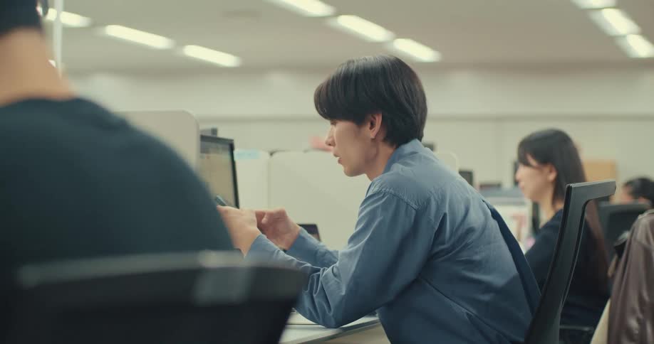 A focused young Asian man works diligently at his desk in a busy office, multitasking between his computer and smartphone