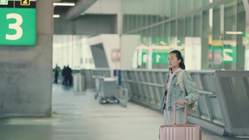An Asian woman with a backpack and pink suitcase walks through a modern airport terminal