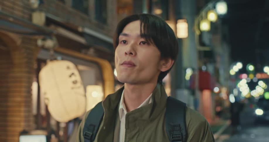 A contemplative young Asian man walks in slow motion down an illuminated city street at night