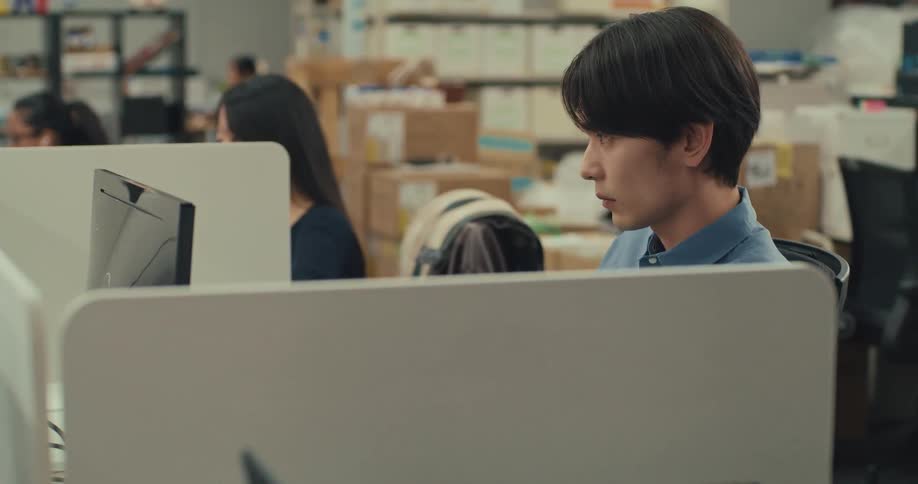 A young Asian man works intently on his computer in a busy office located within a warehouse