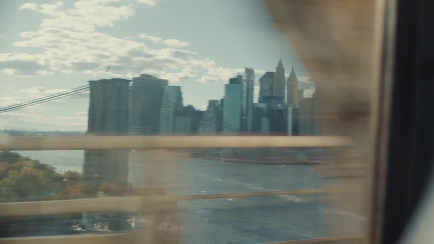 Handheld shot from a moving train window reveals the Brooklyn Bridge and the New York City skyline over the East River