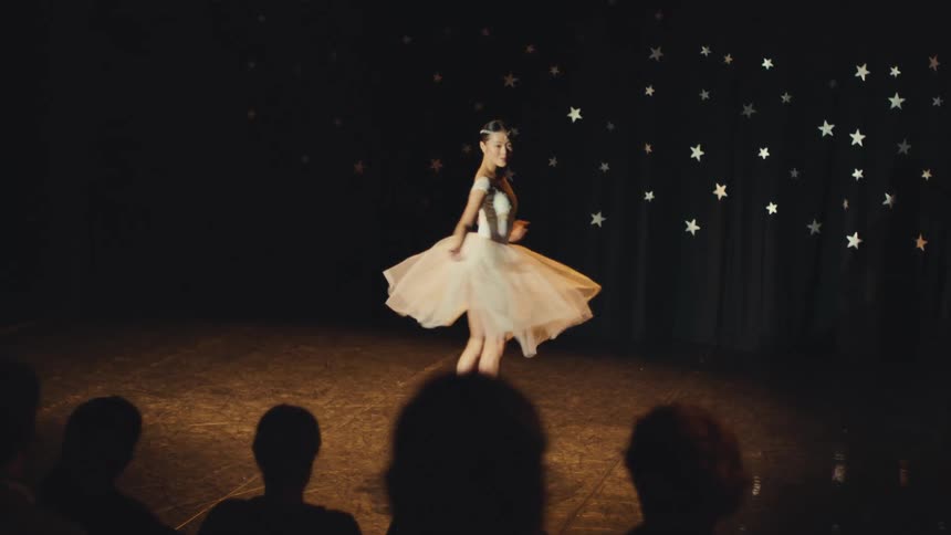 An elegant ballerina dances gracefully under a spotlight on stage in front of a silhouetted audience