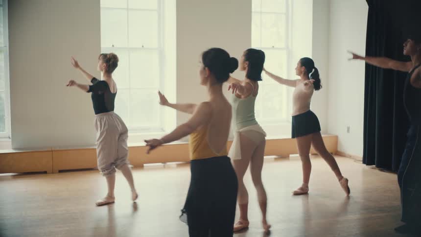 A diverse group of graceful ballet dancers practices choreography together in a bright, sunlit studio in slow motion