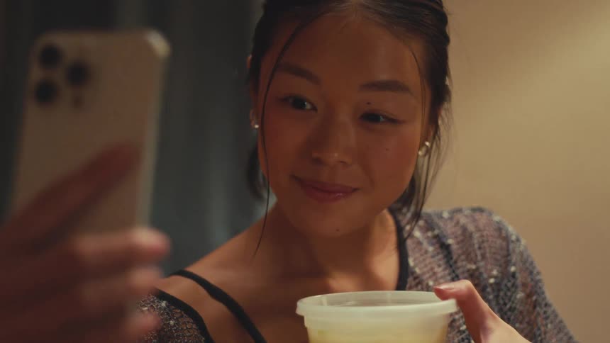 Close up shot of a young Asian woman smiling as she takes a picture of her takeout soup with her smartphone