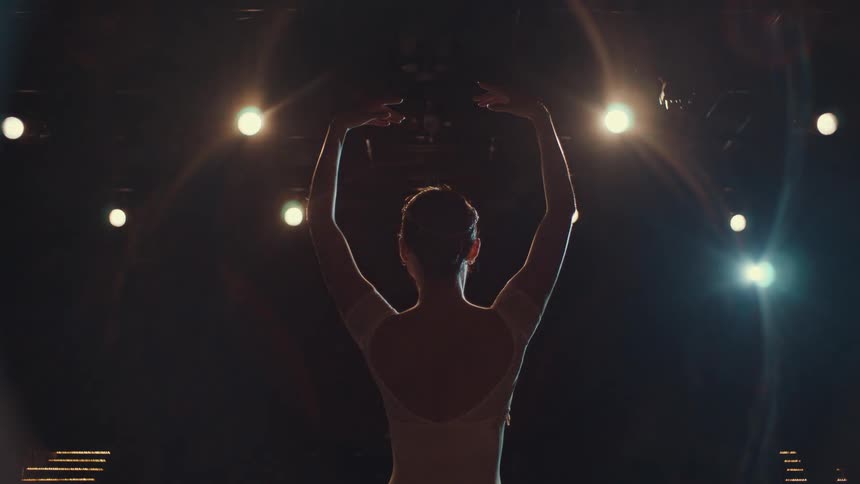A ballerina seen from behind gracefully takes her opening pose on a dark stage as the bright spotlights turn on