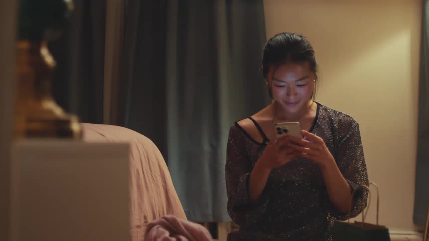 A happy Asian woman in a sparkly top smiles while using her smartphone in her dimly lit bedroom at night