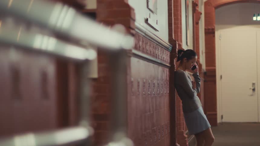 A disheartened woman leans against a tiled wall in a hallway as she listens intently to a phone call