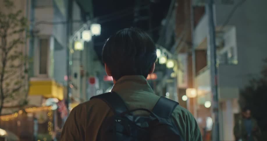 A slow-motion, over-the-shoulder shot follows a young man walking through a vibrant, illuminated city alley at night
