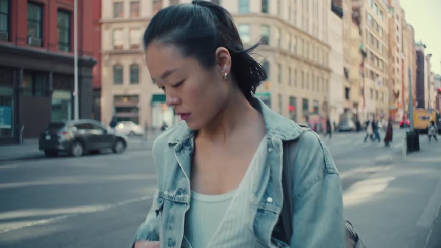 A young Asian woman walking on a bustling city street smiles happily while checking her smartphone