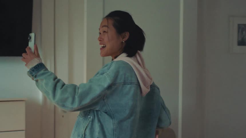 A happy young Asian woman walks into her room and records a video of herself on her smartphone