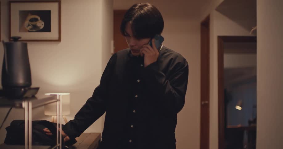 A young Asian man returns home in slow motion, talking on his phone as he walks through the apartment entryway
